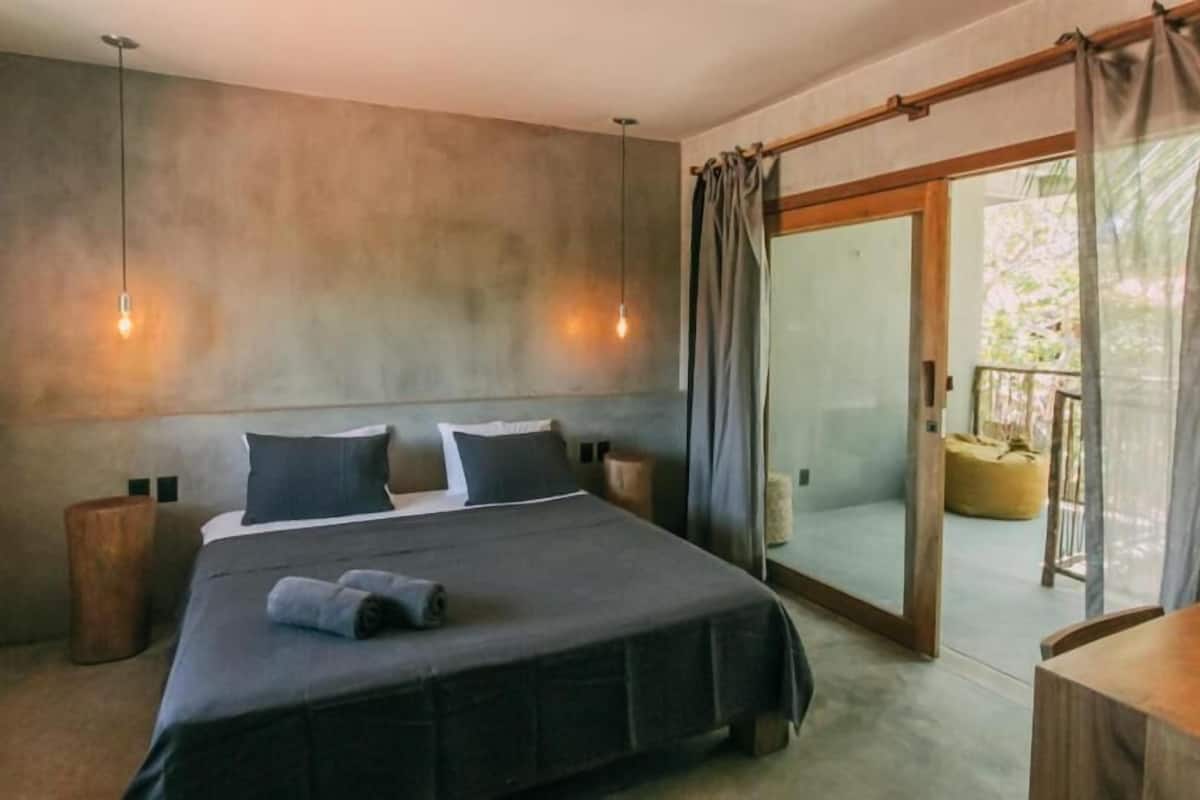 Une chambre dotée d’un grand lit, de deux tables de chevet, d’un banc en bois et d’une porte-fenêtre coulissante donnant sur un espace extérieur.