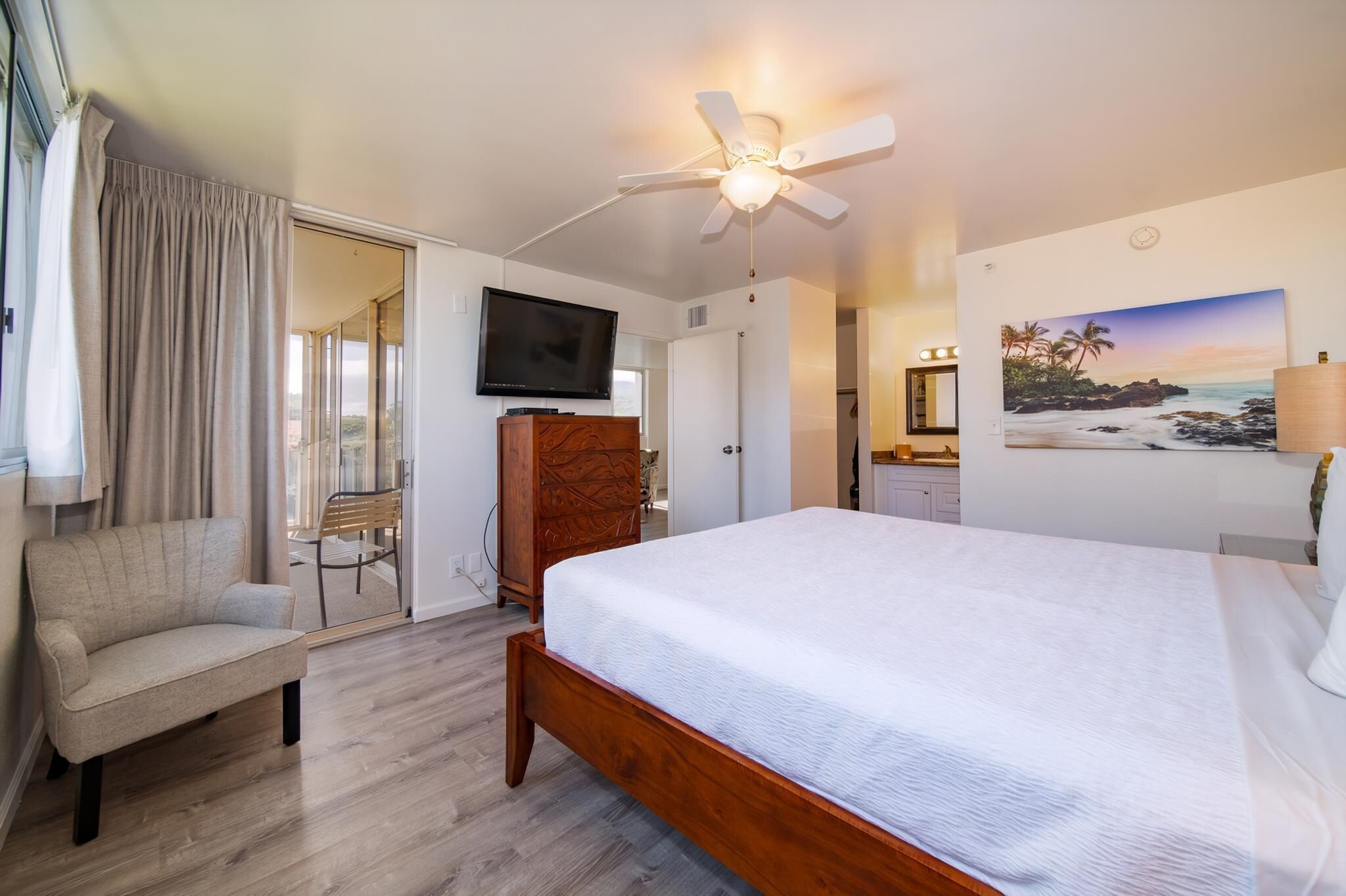 A bedroom with a bed, a chair, a television, a ceiling fan, and a painting on the wall.