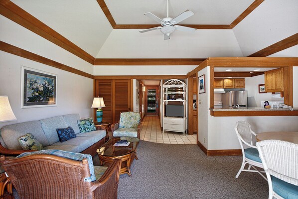 A living room with a sofa, armchairs, a coffee table, a dining area with a table and chairs, a kitchen with a refrigerator, and a ceiling fan.