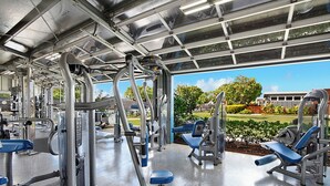 Une salle de sport équipée de divers appareils d’exercice, dotée d’un espace abrité et offrant une vue sur un parc et un immeuble.