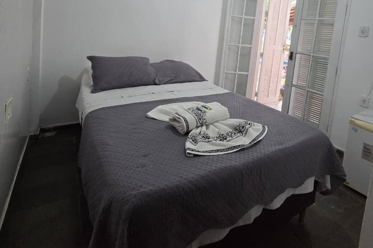 Un lit simple recouvert d’une couverture à motifs gris et blancs, un oreiller blanc et une serviette pliée posés sur le lit. On aperçoit une porte blanche avec des panneaux de verre et un mur blanc avec un interrupteur.