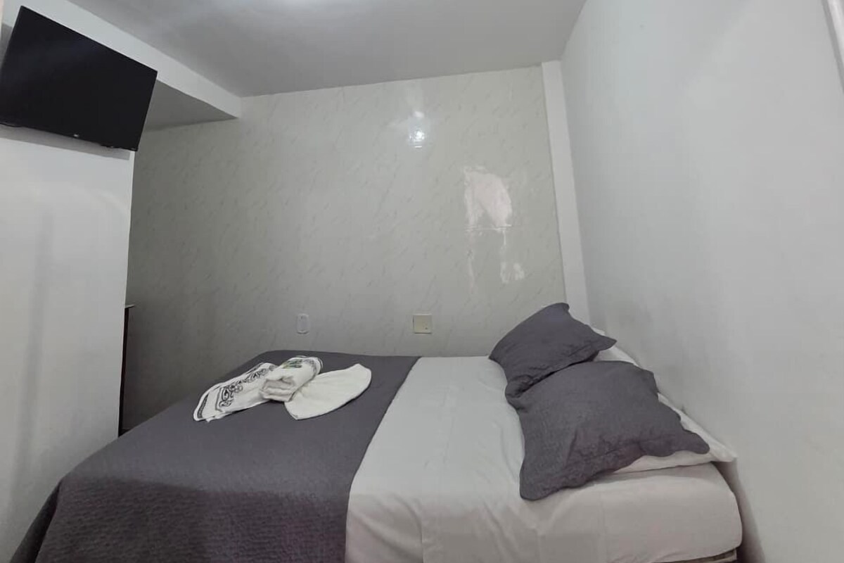 Ein kleines Zimmer mit einem Einzelbett, weißer Decke, einem an der Wand befestigten Flachbildfernseher und einem Fenster auf der rechten Seite.