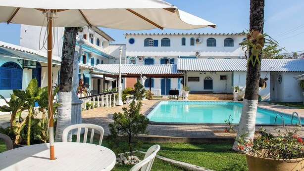 Zona de piscina con una sombrilla blanca, palmeras y un edificio con puertas y ventanas azules.