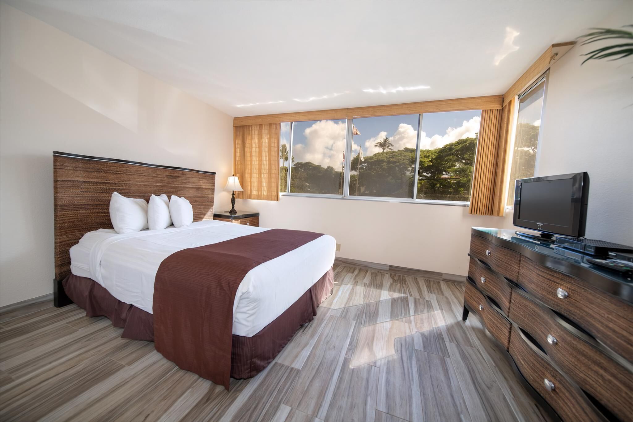 Een hotelkamer met een groot bed, een televisie, een dressoir en een raam met uitzicht op bomen en wolken.
