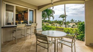 A balcony with a round table and chairs, a kitchen area with cabinets and a window, and a view of a tropical landscape with palm trees and a cloudy sky. - Palms at Wailea Villa 1103 Ocean View 1 BD 2 BA (Wailea)