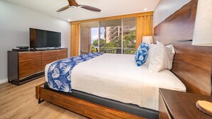 A modern hotel room with a large bed, wooden headboard, a ceiling fan, a TV on a wooden cabinet, and a view of the outside through large windows.