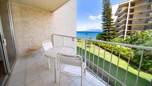A balcony with a table and chairs, a view of a building, and a landscaped garden. - Royal Kahana Maui 303 Ocean View Studio (Lahaina)