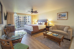 A hotel room with a bed, a sofa, a glass table, a chair, a ceiling fan, and a balcony with a view.