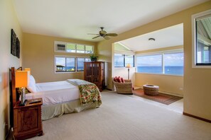 A bedroom with a bed, a nightstand, a lamp, a ceiling fan, a window, a view of the ocean, and a wicker chair.