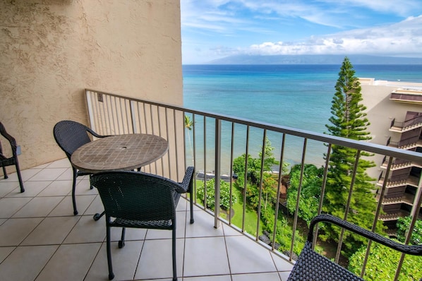 A balcony with a table and chairs, overlooking a coastal view with greenery and a distant mountain.