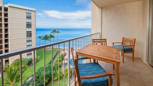 Un balcon avec une table et des chaises en bois, donnant sur un océan d’un bleu limpide et sur une zone verdoyante parsemée de palmiers.