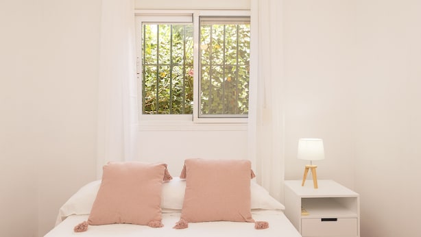 A bedroom with a bed, two pillows, a nightstand with a lamp, and a window with greenery outside. - El Cortijo in La Linea de la Conception (La Línea de la Concepción)