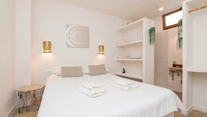 A neatly made bed with towels, a bedside table, wall-mounted shelves, and a hanging plant. - Pura Vida in Vejer de la Frontera (Vejer de la Frontera)