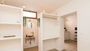 A compact, well-lit room with a white wall, a wooden floor, a small sink with a brass faucet, a hanging plant, and a clothes drying rack. - Pura Vida in Vejer de la Frontera (Vejer de la Frontera)