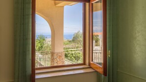 A room with a large window, a balcony with a stone pillar, and a view of the sea and greenery. - Villa With Volcano View (Zafferana Etnea)