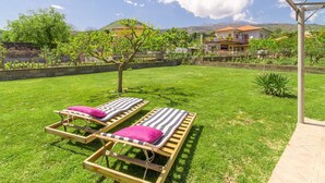A backyard with two wooden sun loungers, a small tree, and a house in the background. - Villa With Volcano View (Zafferana Etnea)