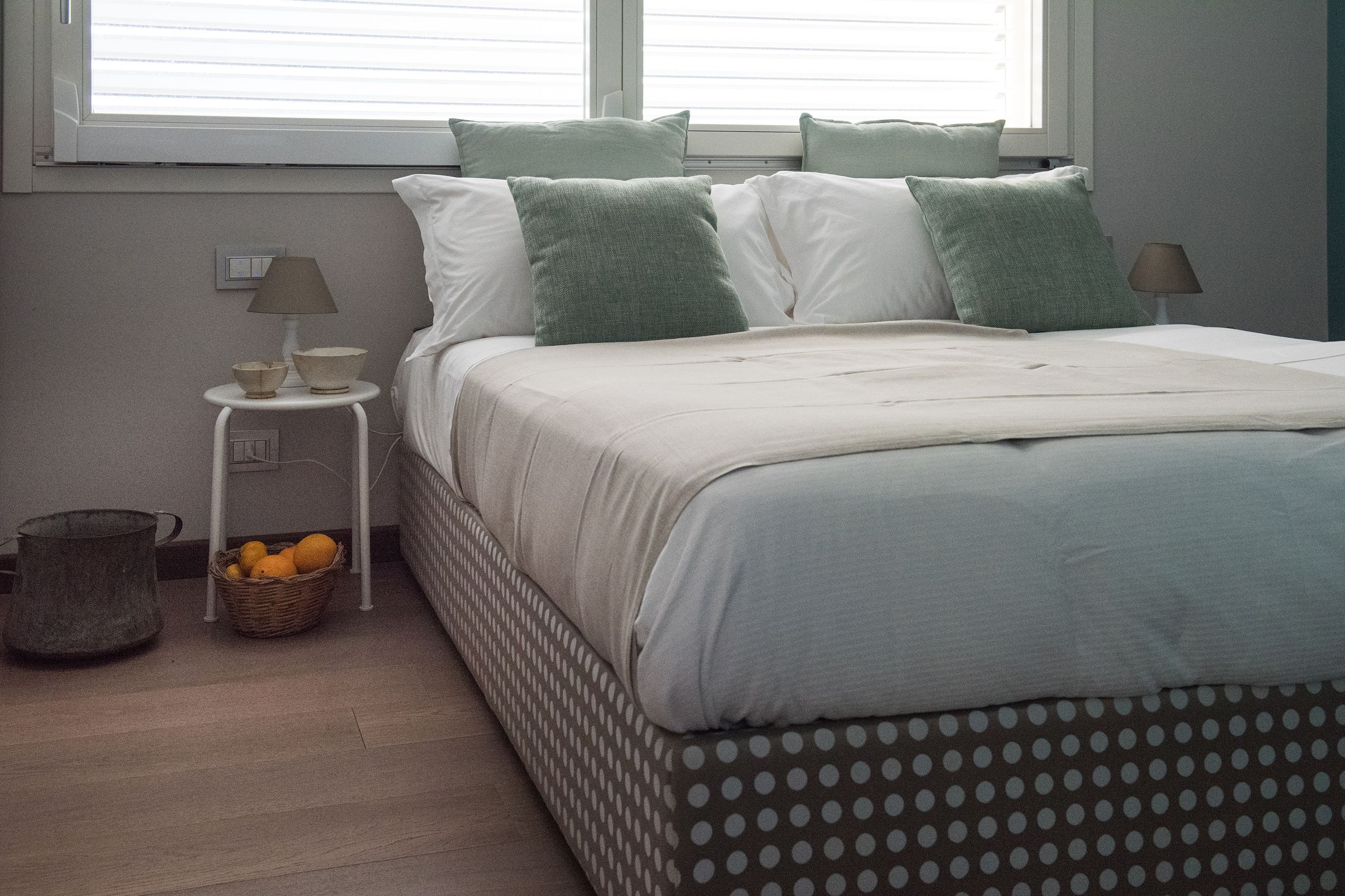 A neatly made bed with a polka dot bedspread, a bedside table with a lamp, a basket of oranges, and a window with blinds.
