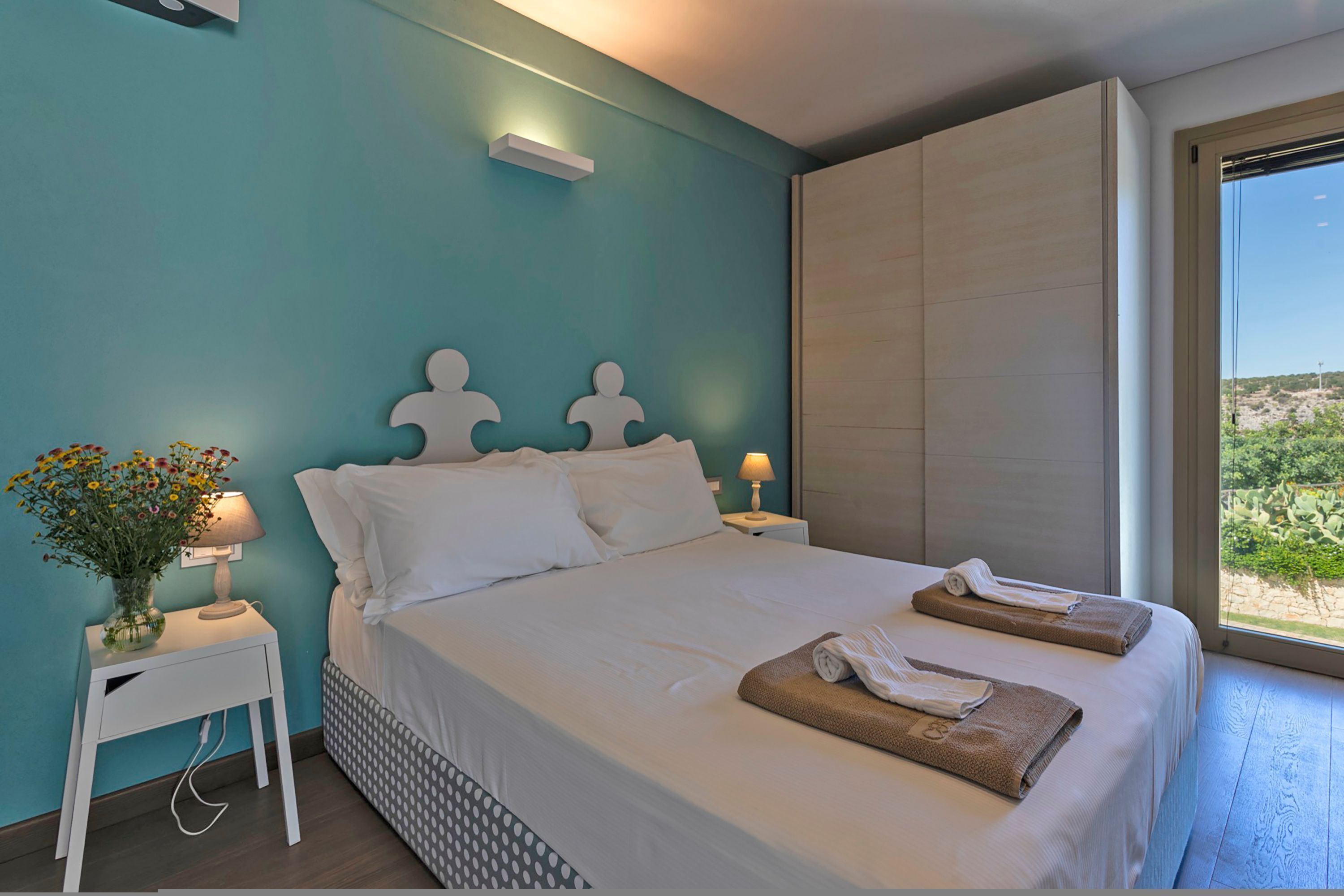 A bedroom with a bed, white nightstands, a vase with flowers, and a window with a view.