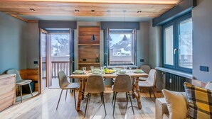 A dining area with a wooden table set for six, surrounded by chairs, with a view of mountains through large windows. - Simona Luxuryapartment few Steps From ski (Sestriere)
