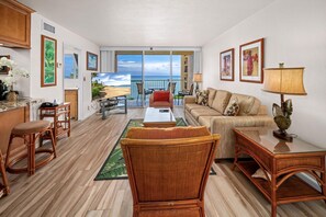 A spacious living room with a large window overlooking a beach, furnished with wicker chairs, a sofa, and a coffee table.
