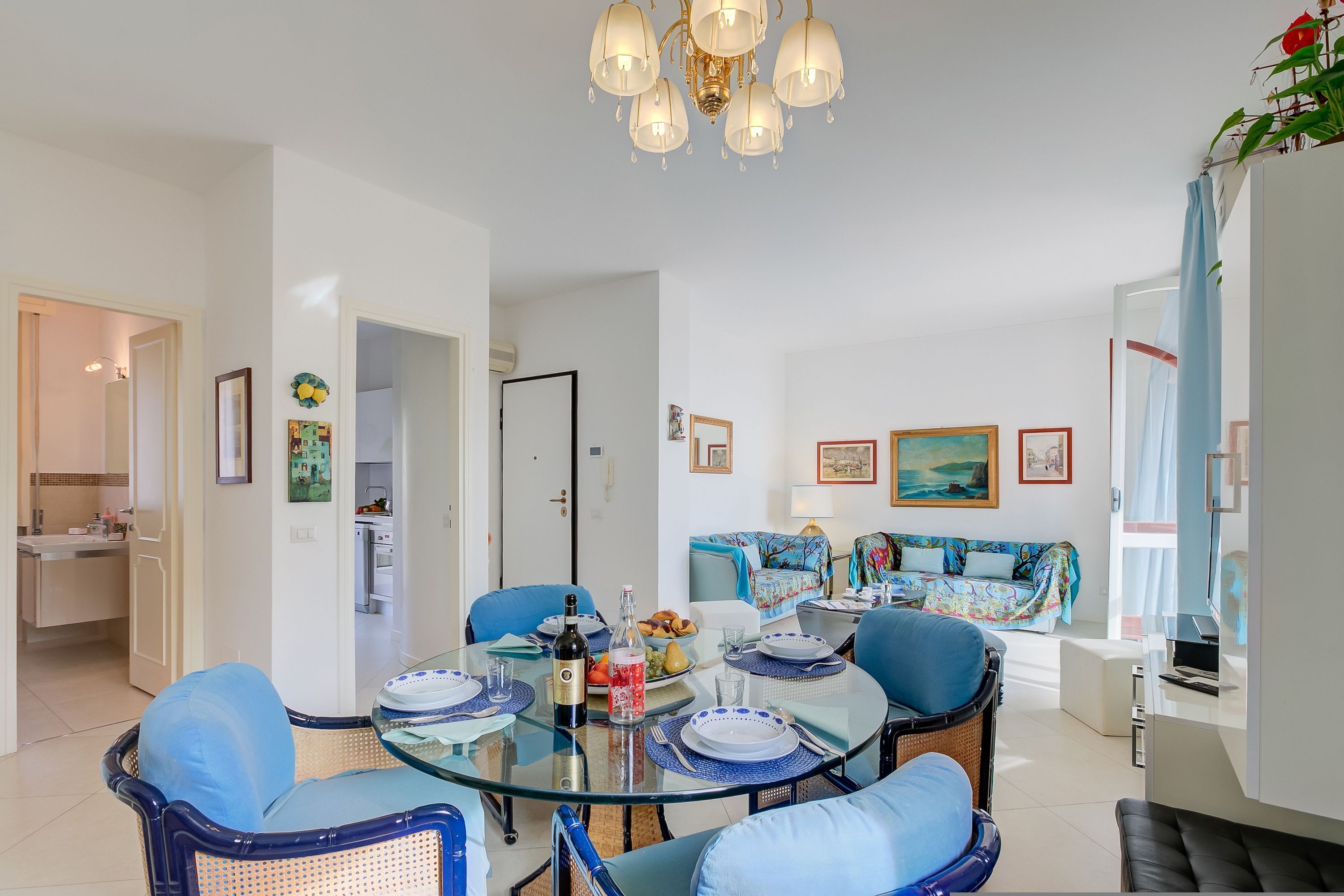 A modern living room with a glass dining table, blue chairs, a sofa, and a chandelier.