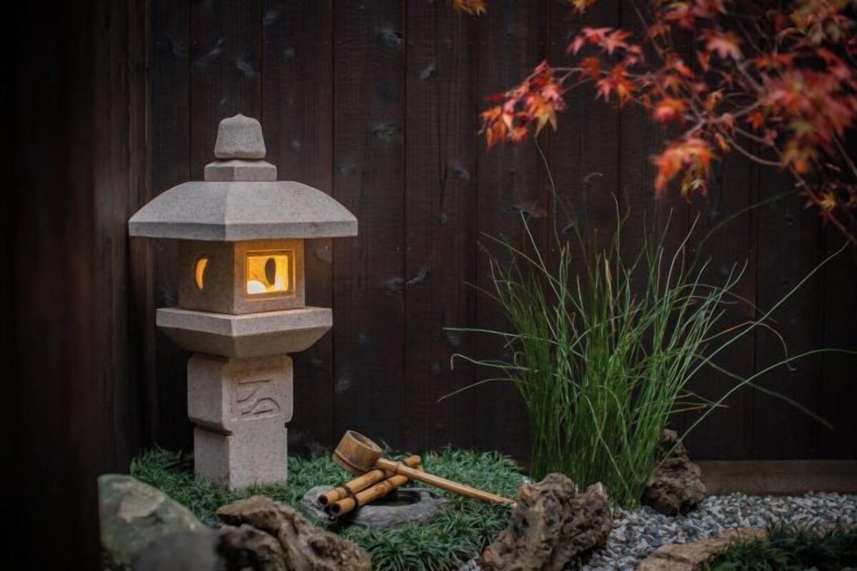A stone lantern with a lit interior, placed on a bed of pebbles with a wooden mallet beside it, and a plant with red leaves in the background.