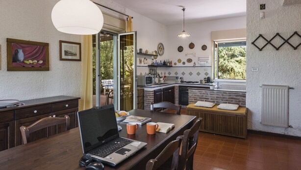 A dining area with a laptop on the table, a bench, and a kitchen in the background. - Pedara Escape With Garden by Wonderful Italy (Pedara)