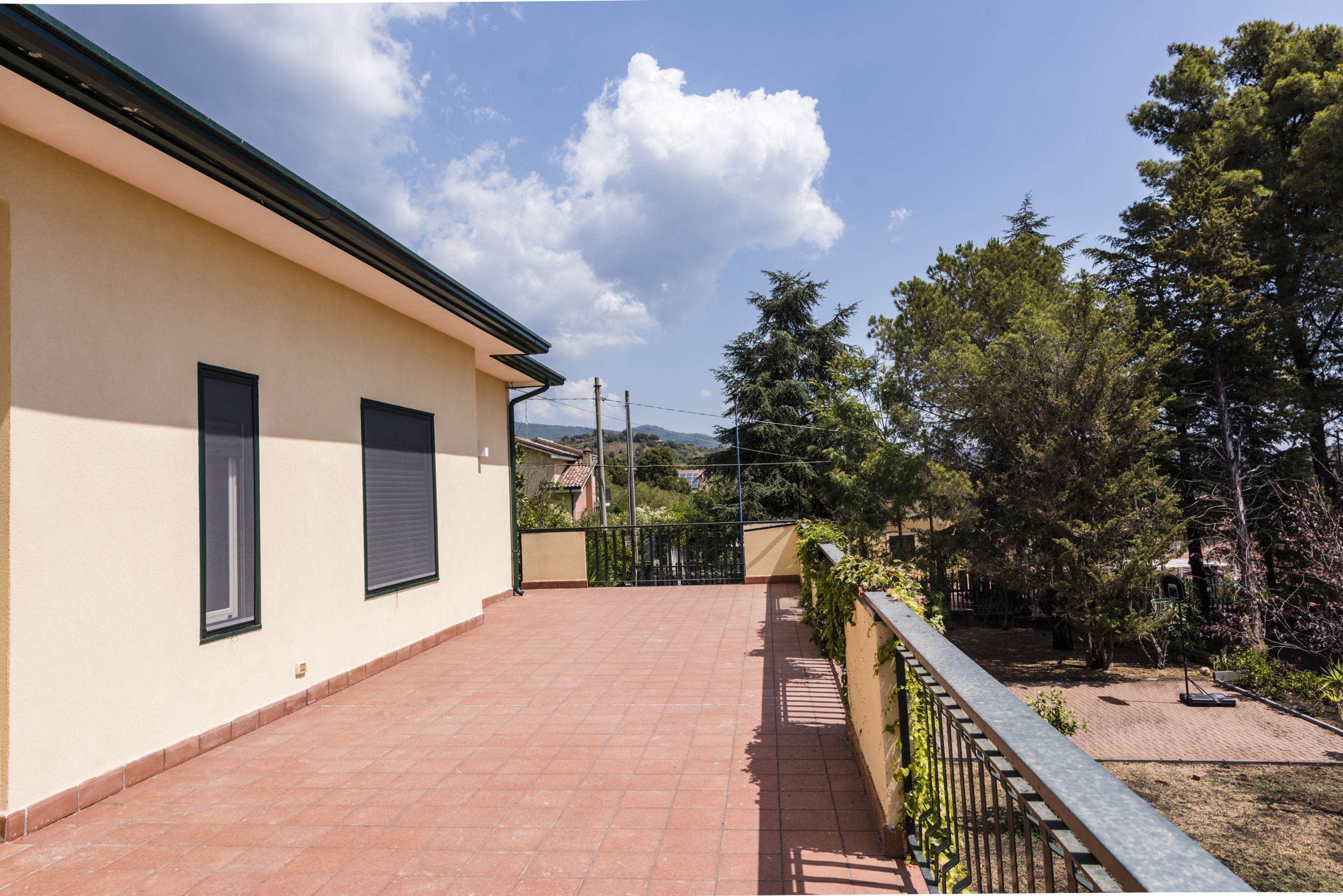 A building with a tiled patio, a balcony with a metal railing, and a view of trees and hills in the background.