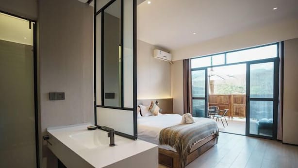 A modern bedroom with a large bed, a bathroom with a white sink, and a sliding glass door leading to an outdoor area. - Nanlahe Paya Resort Hotel (Mengla)