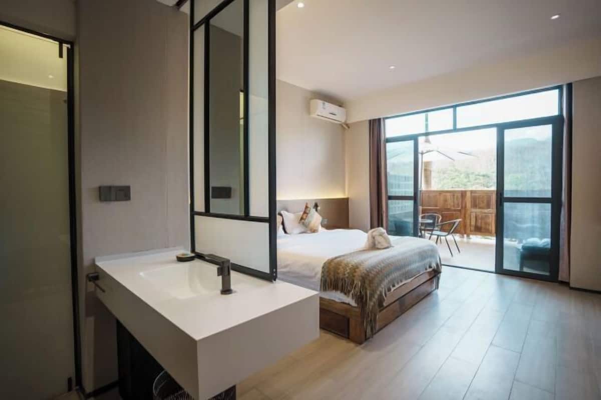 A modern bedroom with a large bed, a bathroom with a white sink, and a sliding glass door leading to an outdoor area.