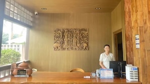 A reception area with a wooden wall, a large decorative panel, a receptionist, and a reception desk with a computer and some items on it. - Nanlahe Paya Resort Hotel (Mengla)