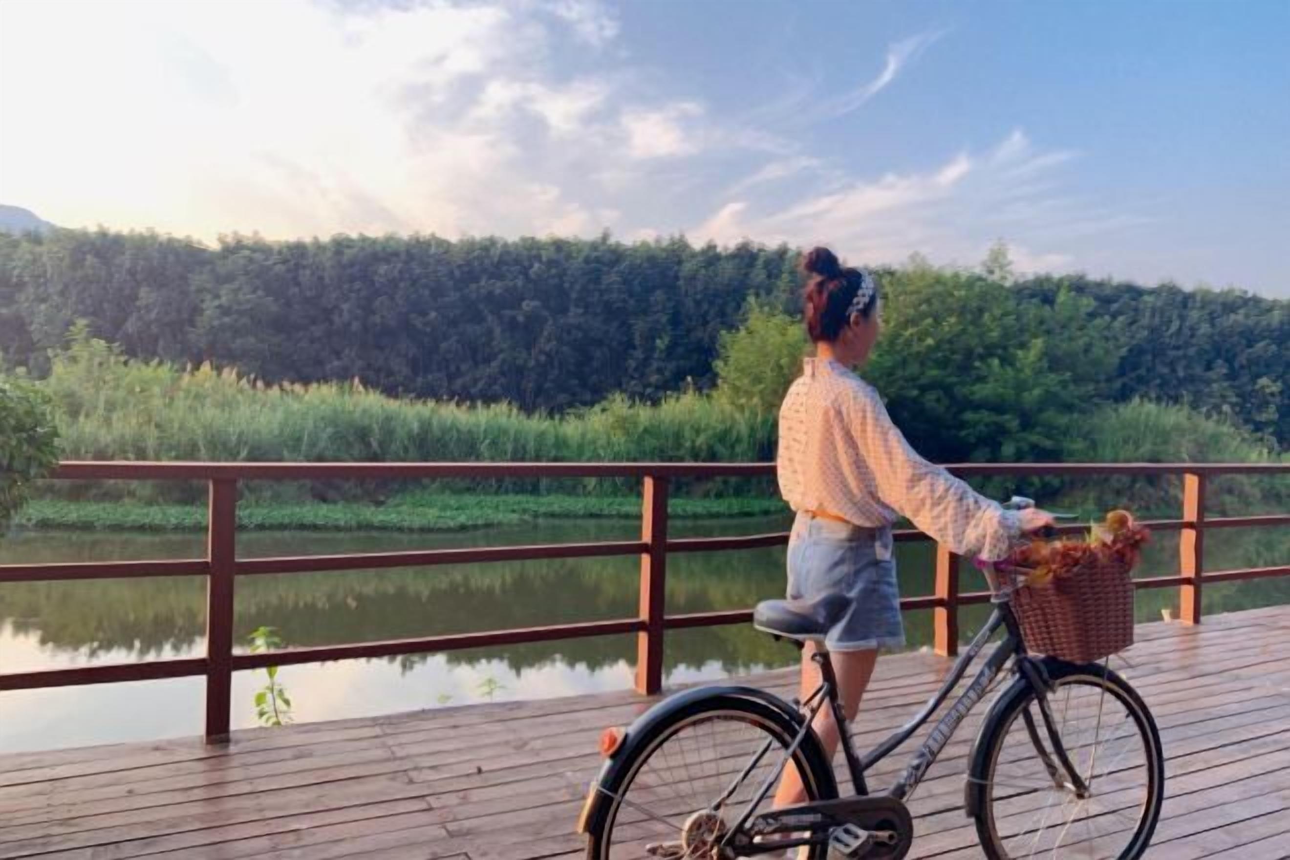 A person on a wooden deck with a bicycle, overlooking a calm body of water and dense greenery.