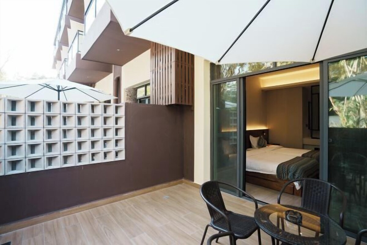 A modern hotel room with a large sliding glass door leading to a balcony, a bed with a nightstand, and a seating area with two chairs and a table.