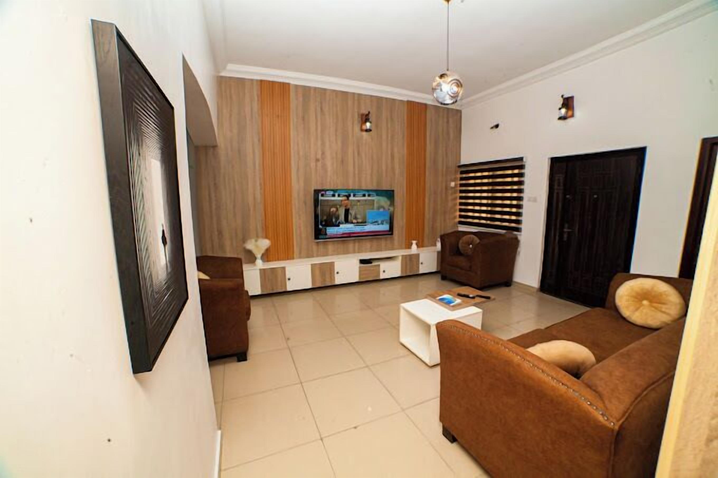 A modern living room with a flat-screen TV mounted on a wooden wall, a brown sofa, a white coffee table, and a brown armchair.