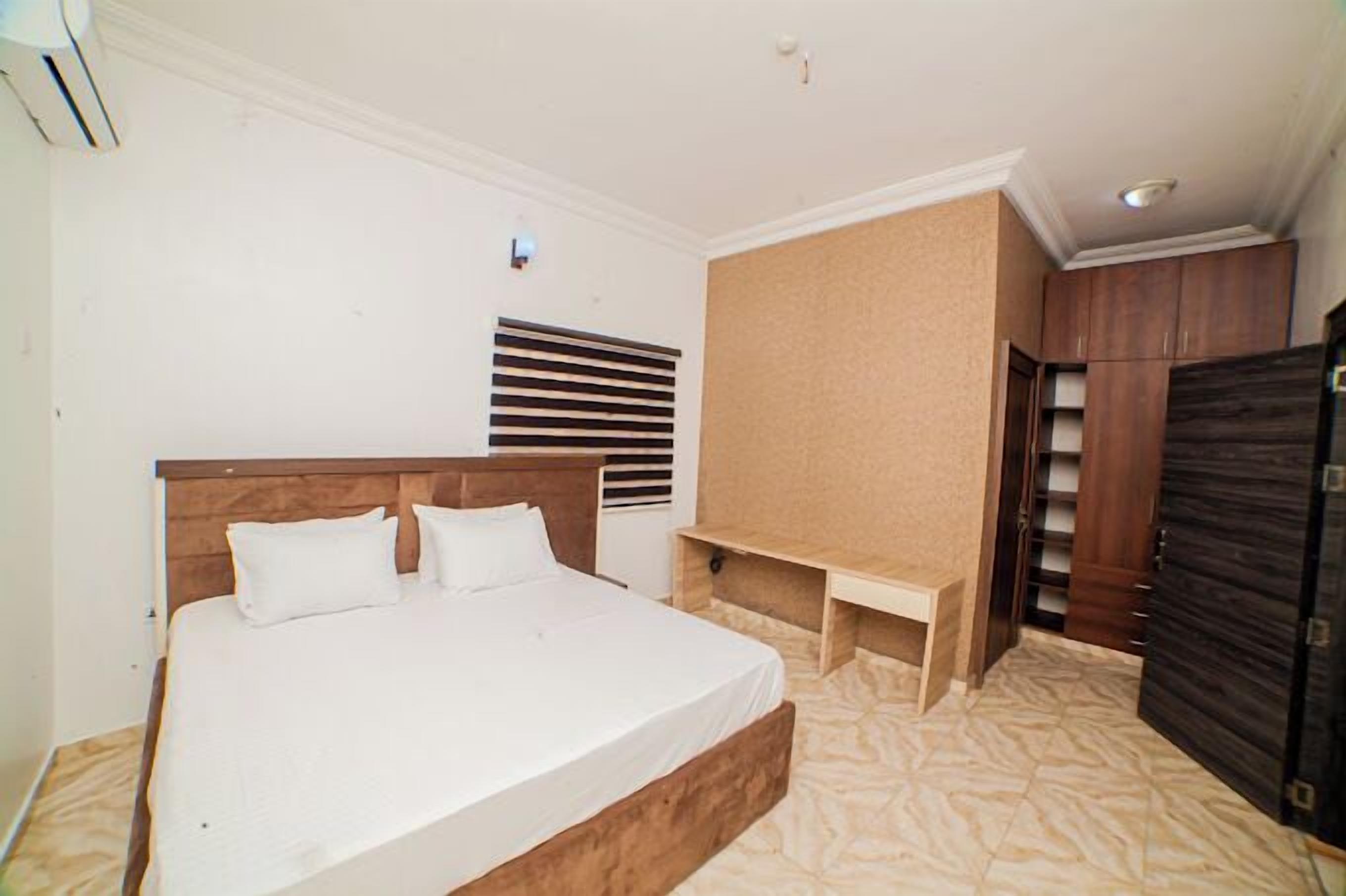 A bedroom with a large bed, a wooden headboard, a built-in bench, a wardrobe, and a window with blinds.