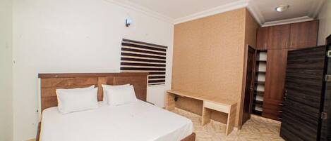 A bedroom with a large bed, a wooden headboard, a built-in bench, a wardrobe, and a window with blinds.