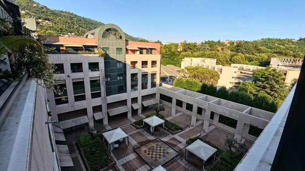 A courtyard with a modern building featuring a curved glass facade, surrounded by a low wall and small tables and chairs. - Como Apartment + Free parking & WiFi (Como)