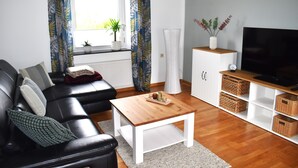 Un salon avec un canapé en cuir noir, une table basse en bois, un meuble blanc avec des paniers et un téléviseur posé sur un meuble en bois.