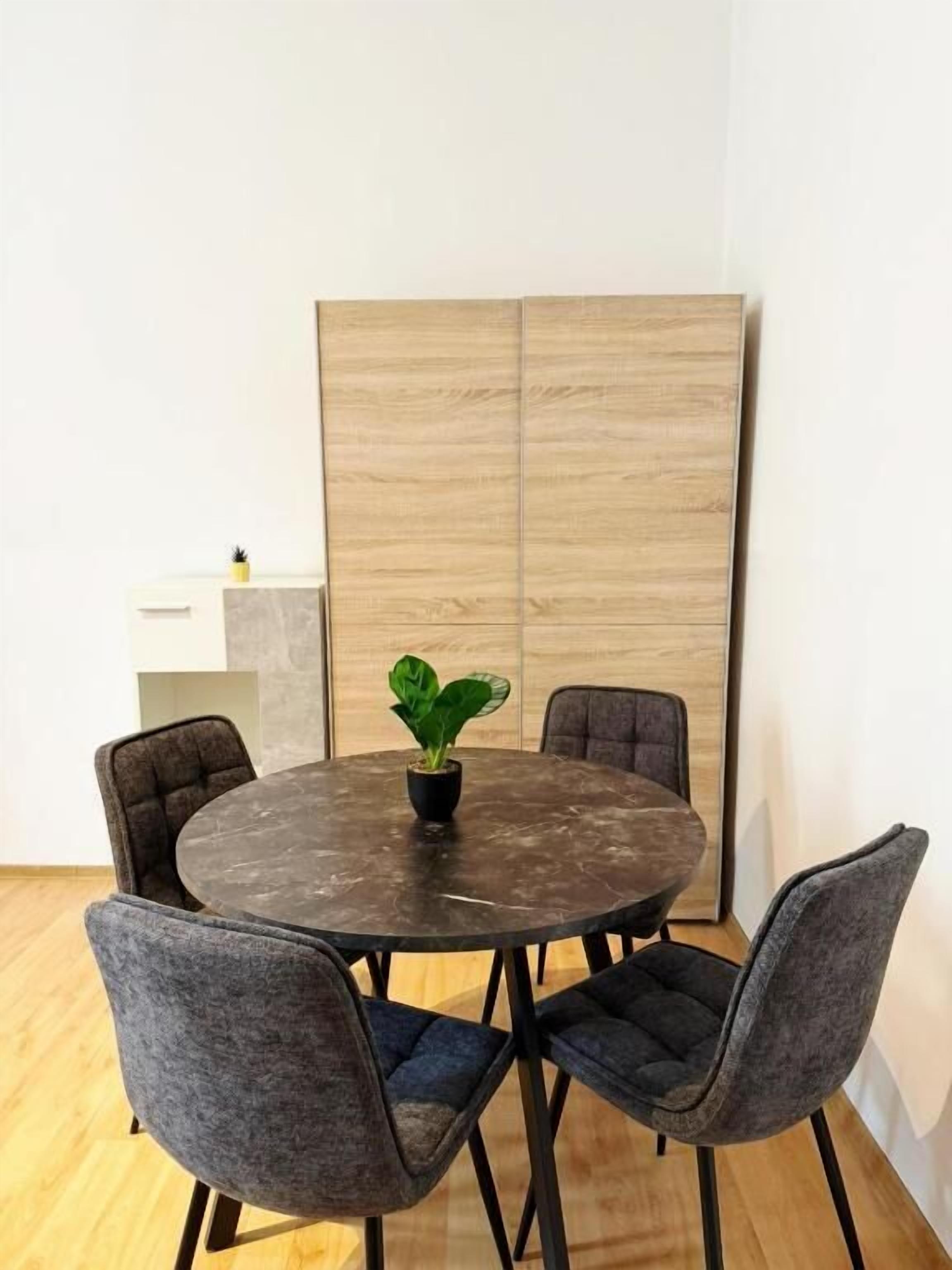 Mesa redonda con cuatro sillas, una planta en maceta y un panel de madera en una habitación con pared blanca y suelo de madera.