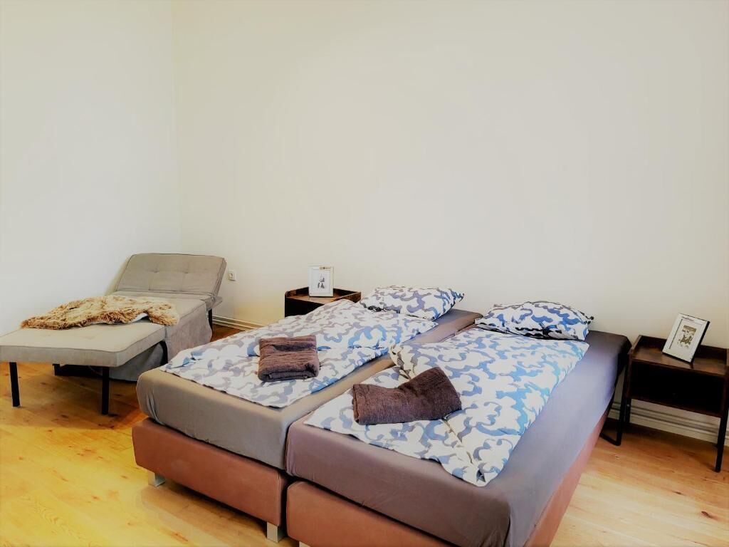 Habitación individual con una cama, un sofá gris, una mesita de noche con una lámpara y un cuadro enmarcado en la pared.