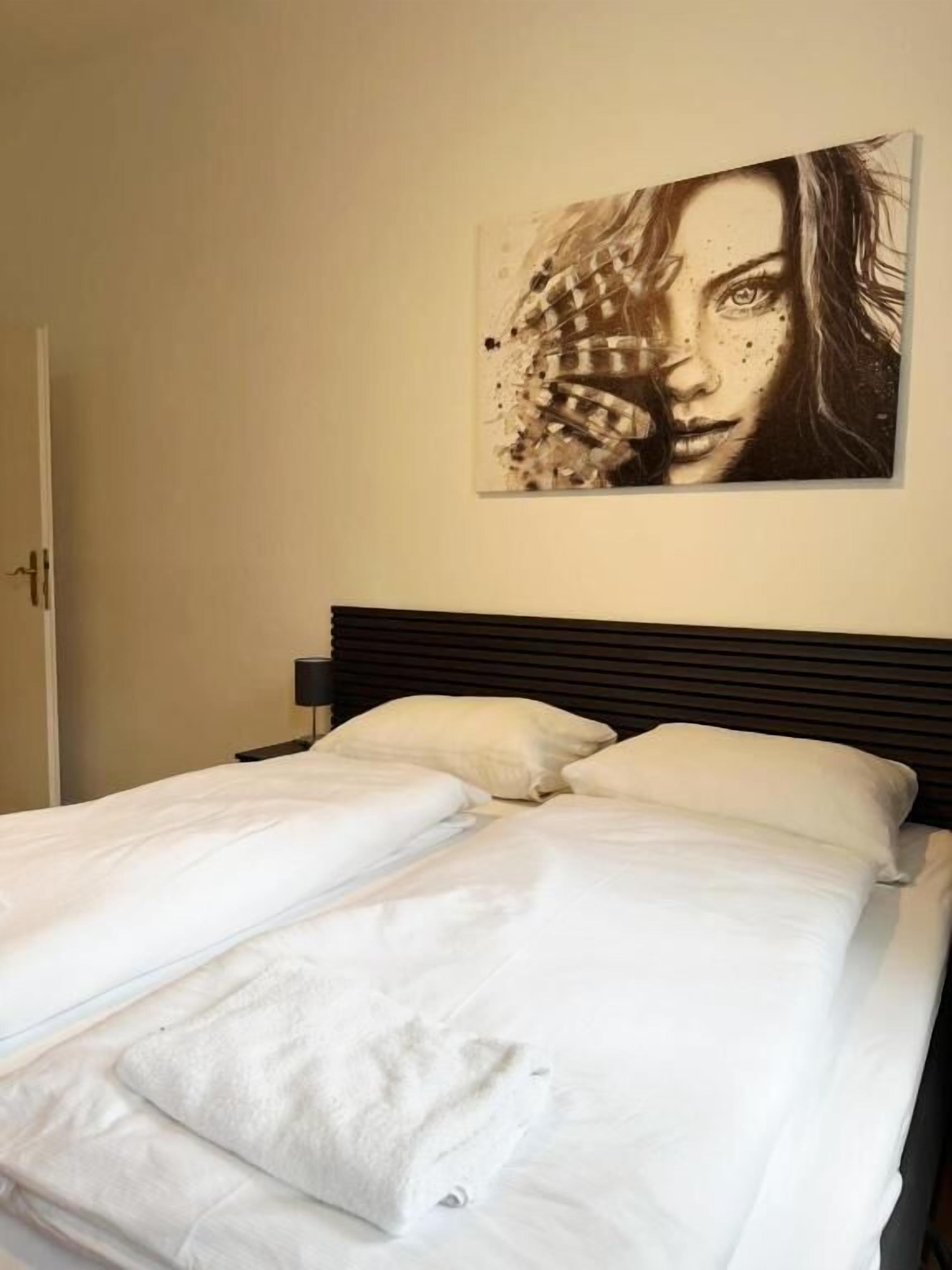 A bed with white bedding and a dark headboard, a wall-mounted artwork, and a door to the left.