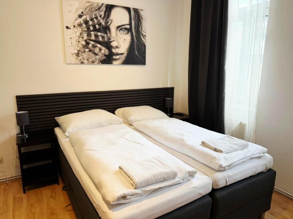 Una cama doble con ropa de cama blanca y un cabecero negro, una mesita de noche con lámpara, y una obra de arte colgada en la pared.
