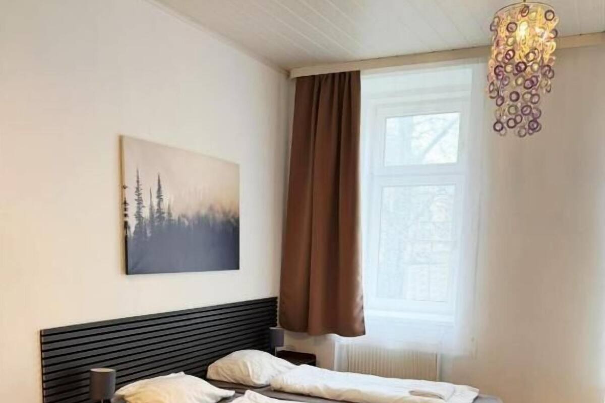Een slaapkamer met een bed, nachtkastje, lamp, schilderij, raam met gordijnen en een hanglamp.