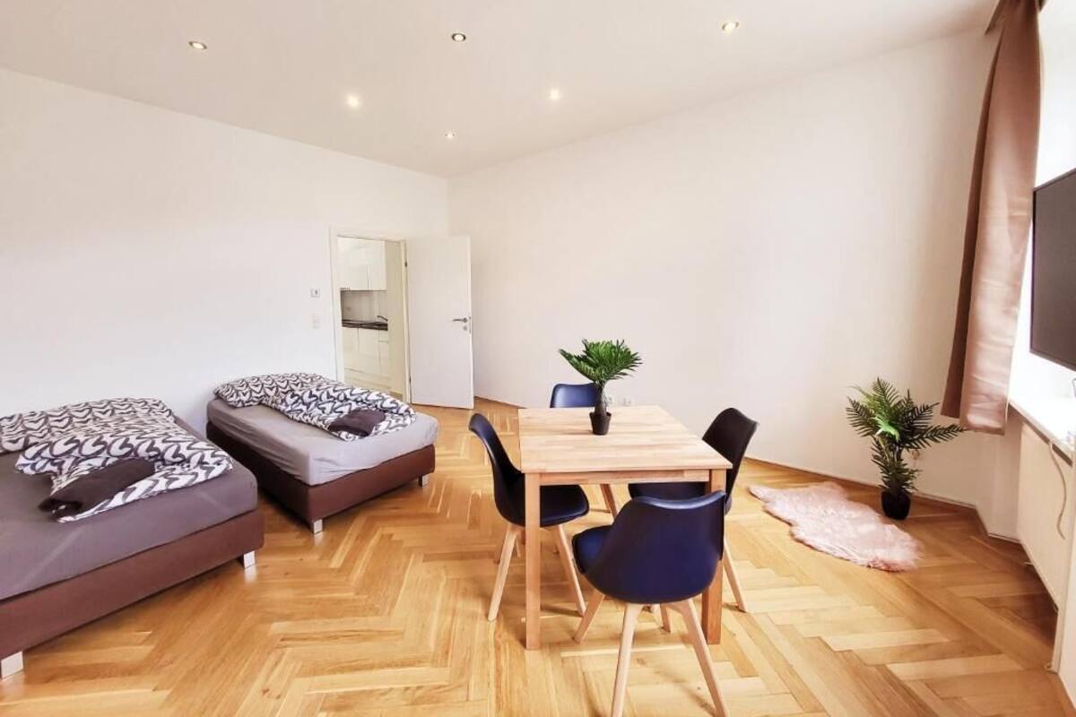 Een moderne kamer met houten vloer, een eettafel met stoelen, twee bedden, een kleine plant en een televisie.