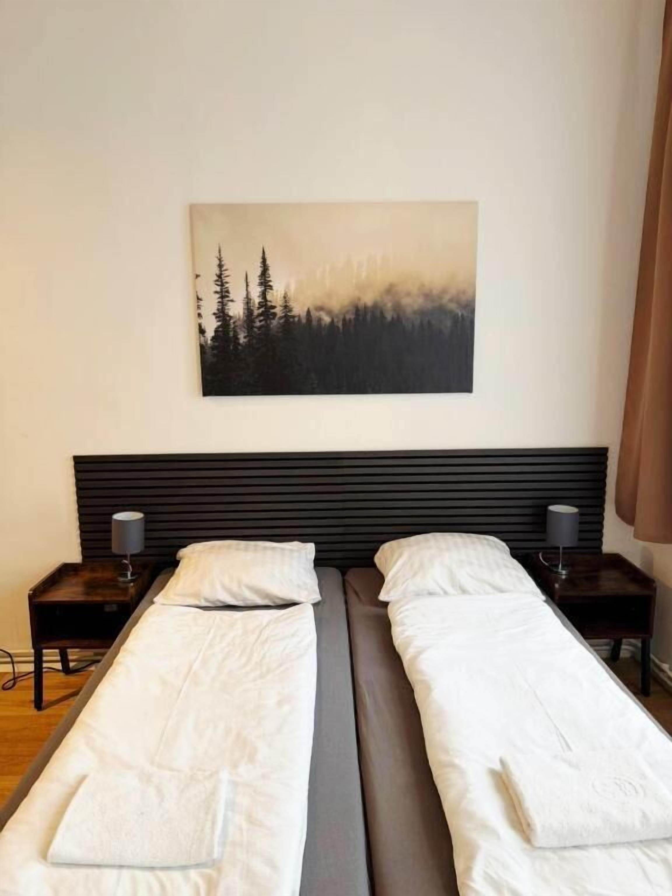 Una cama doble con cabecera, dos mesitas de noche con lámparas y un cuadro de un bosque en la pared.