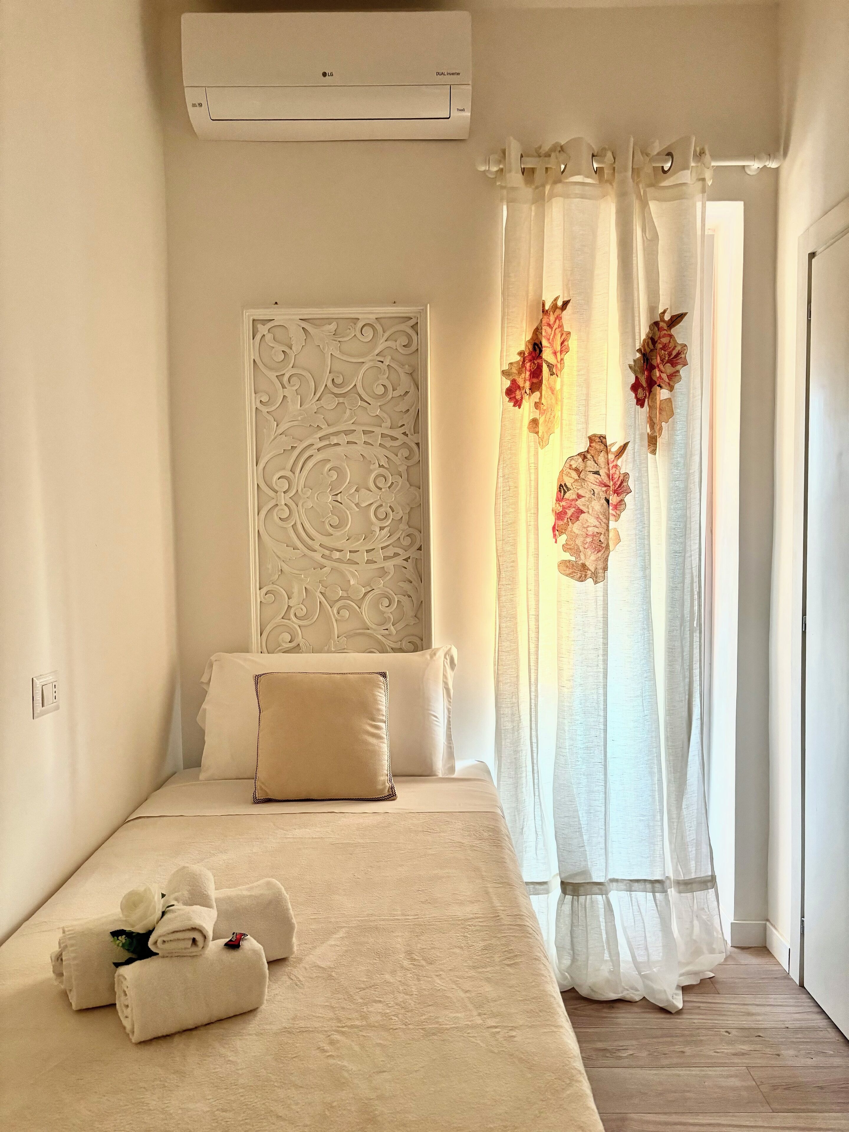 A bedroom with a bed, a white air conditioning unit, a decorative headboard, sheer curtains with floral decorations, and neatly folded towels on the bed.