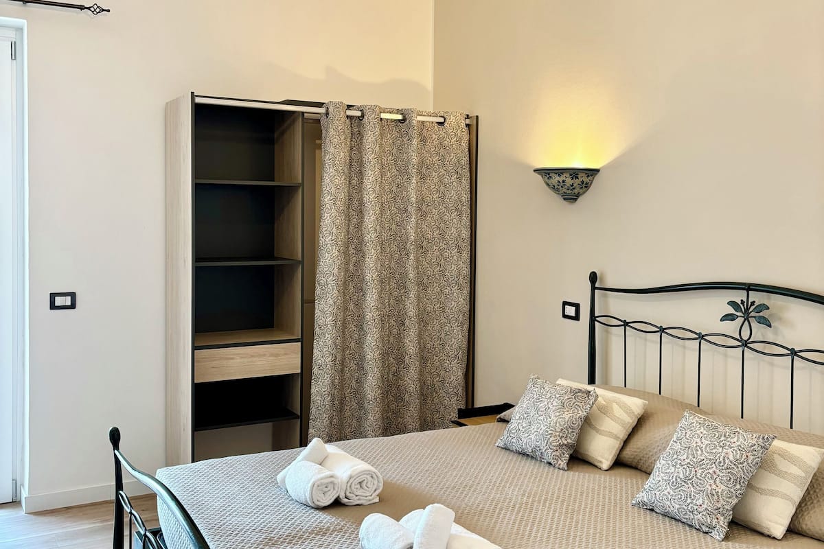 A neatly made bed with rolled towels, a wardrobe, a shower curtain, and a wall-mounted light fixture.
