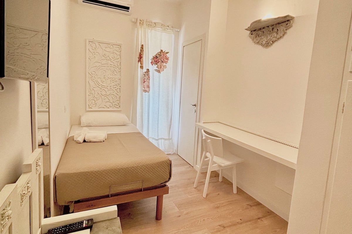 A compact bedroom with a bed, a desk, and a chair. There is a mirror, a wall-mounted air conditioner, and a decorative wall piece.