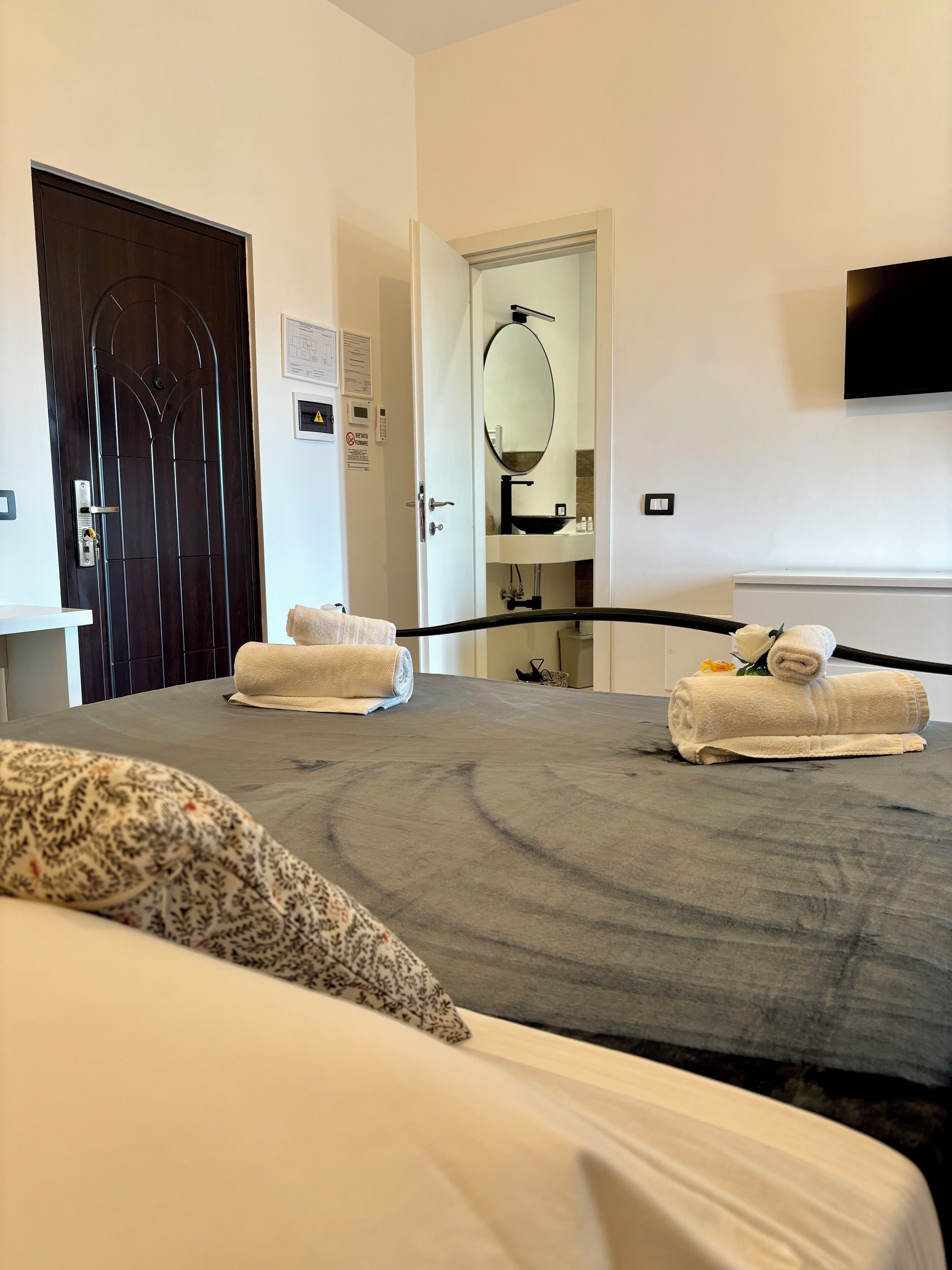 Ein Hotelzimmer mit einem großen Bett, zwei aufgerollten Handtüchern, einer dunklen Holztür, einer weißen Wand mit einem angebrachten Fernseher und einem durch eine geöffnete Tür einsehbaren Badezimmer.