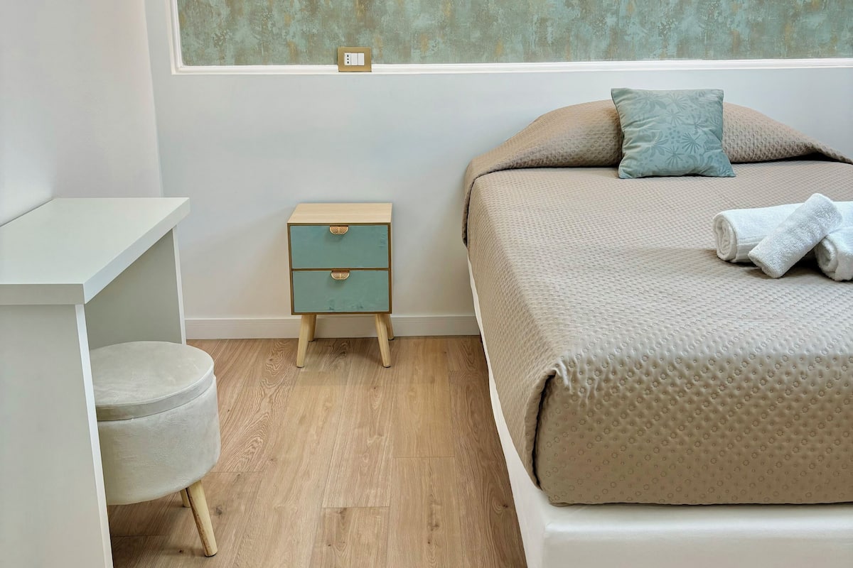 A modern bedroom with a bed, bedside table, nightstand, and a window with a textured wall.